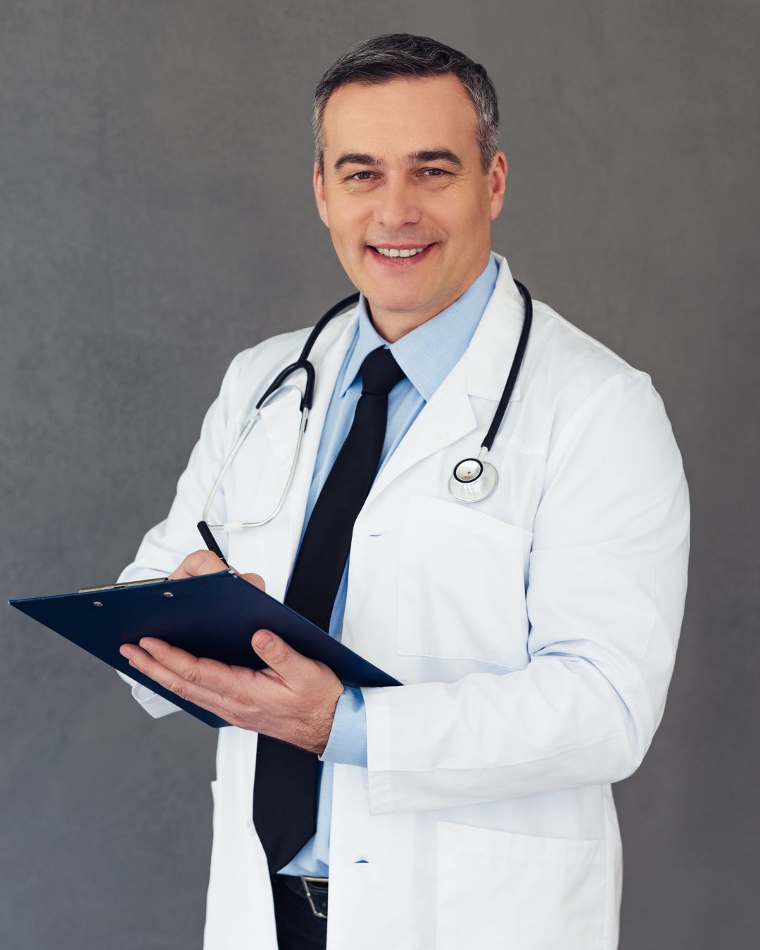 Tell me about your habits! Mature male doctor making notes in clipboard and looking at camera with smile while standing against grey background